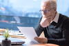 an older man wearing glasses sits at a table looking at a page in front of his laptop